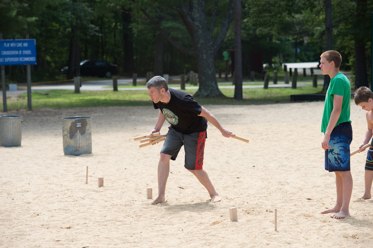 A man prepares to throw wooden sticks in a beach game while two boys watch nearby.