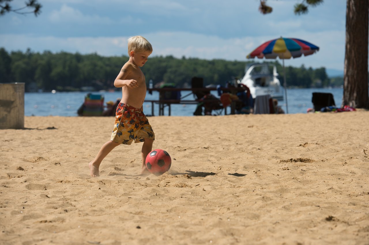A young boy in swim trunks kicks a red soccer ball on a sandy beach near the lake.