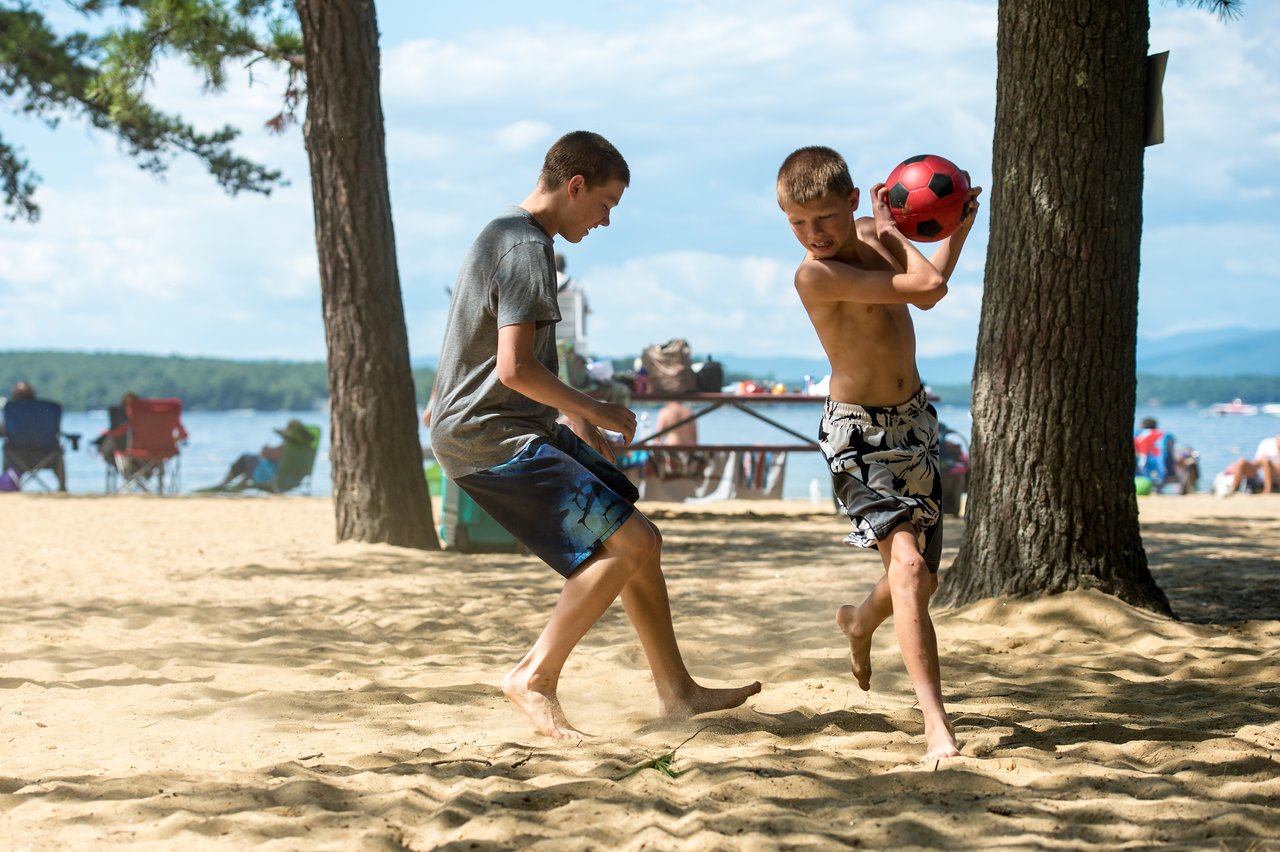Two boys play with a red ball on a sandy beach near Lake Winnipesaukee, surrounded by trees and people.