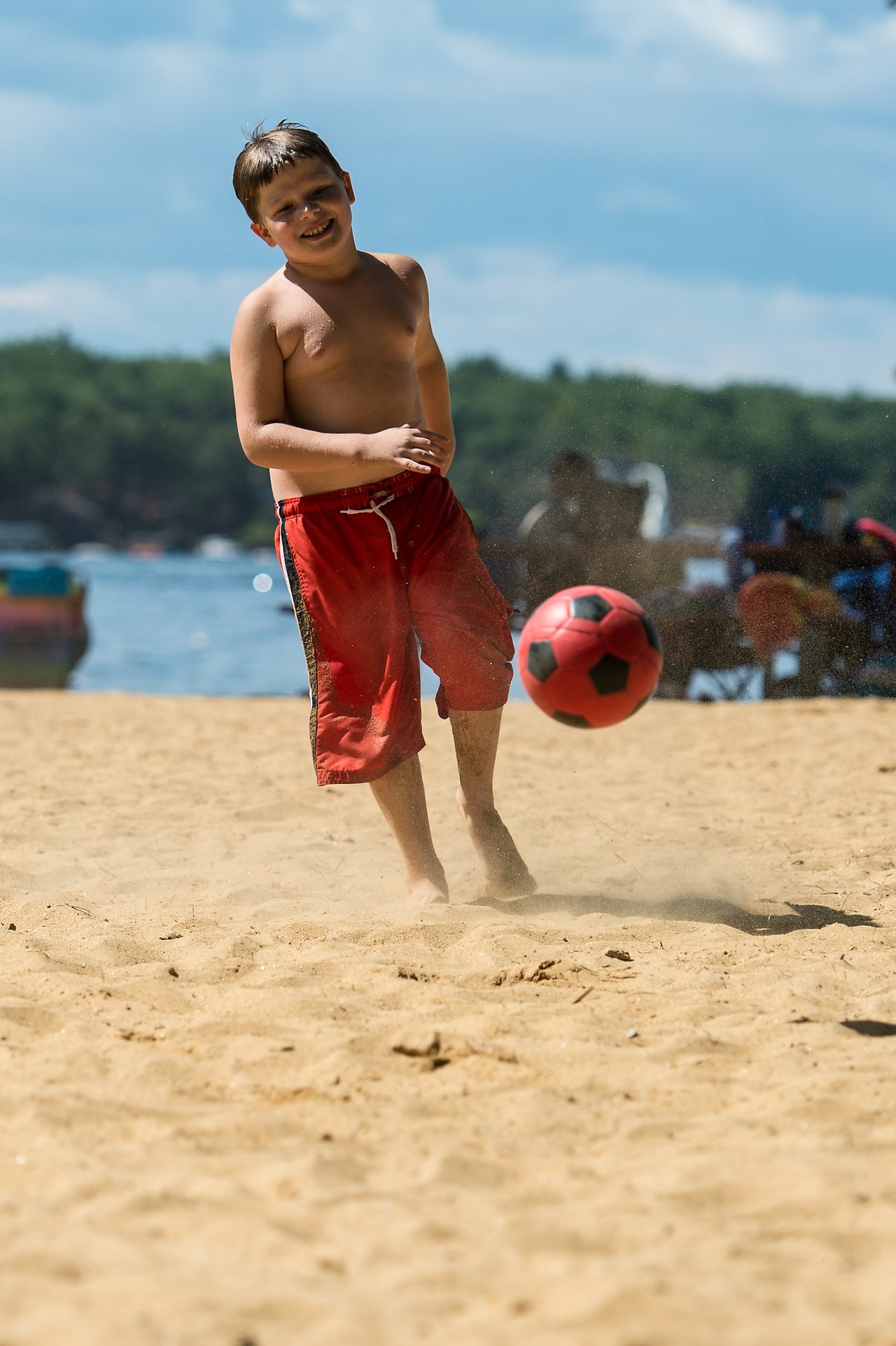 A boy in red shorts kicks a soccer ball on a sandy beach near the water.