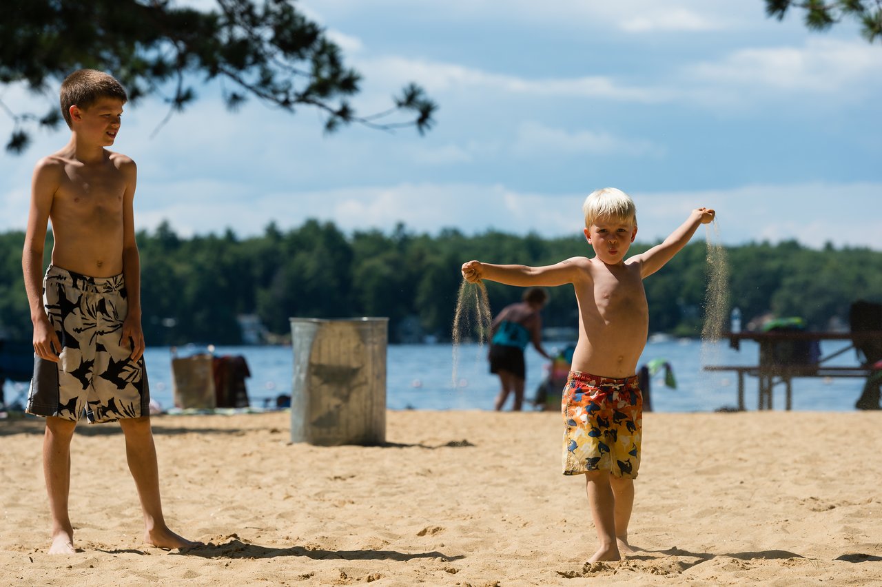 Two children play on a sandy beach near a lake, one tossing sand while the other watches.