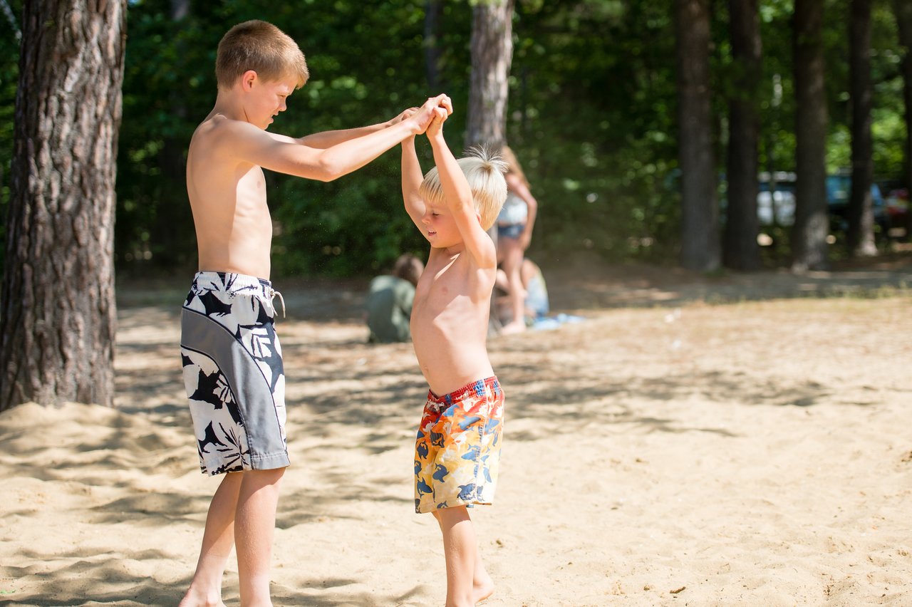 Two children in swim trunks play on a sandy beach, holding hands and smiling under the sunlight.