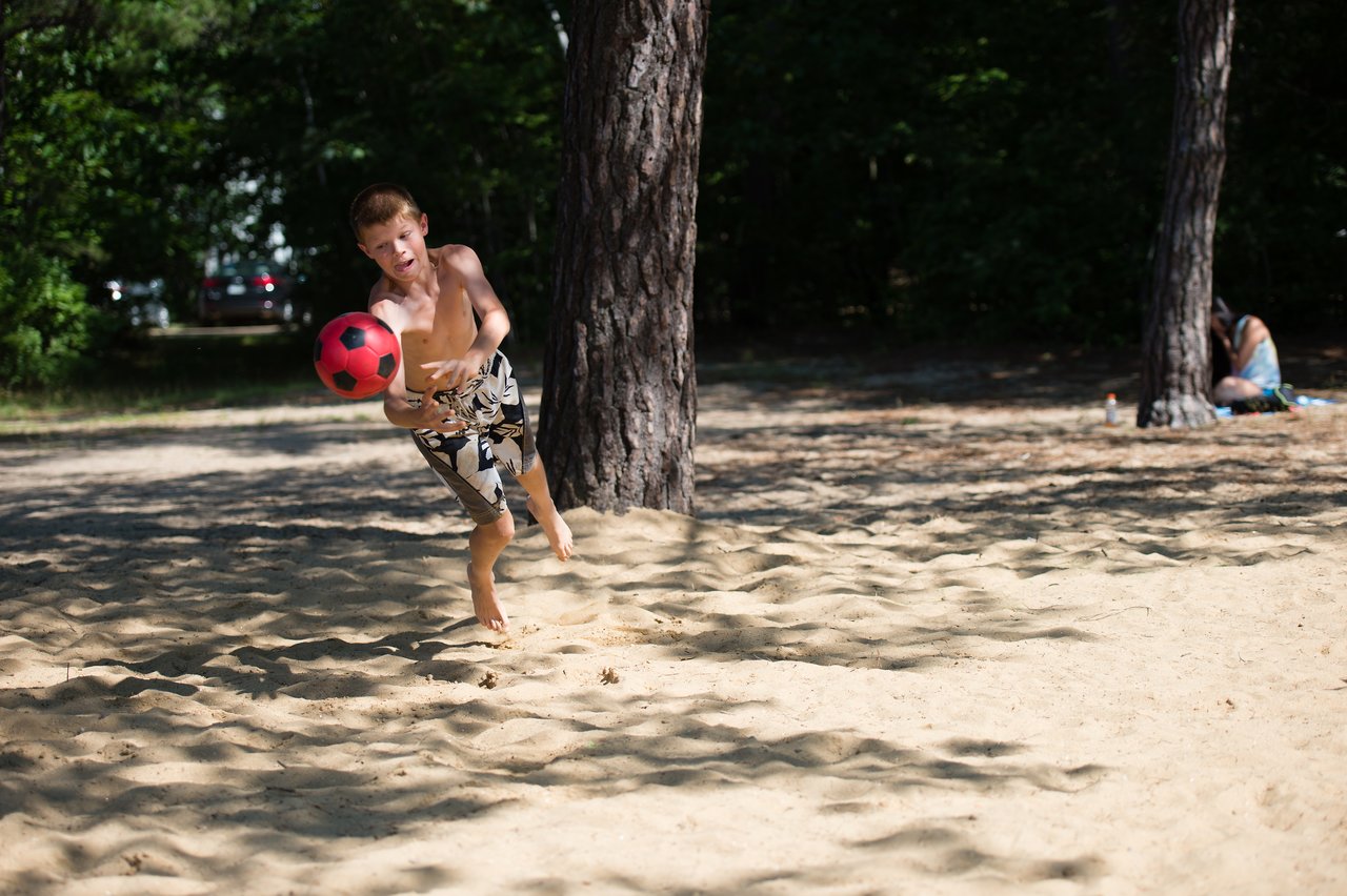 A boy in swim trunks throws a red soccer ball while playing on a sandy beach.