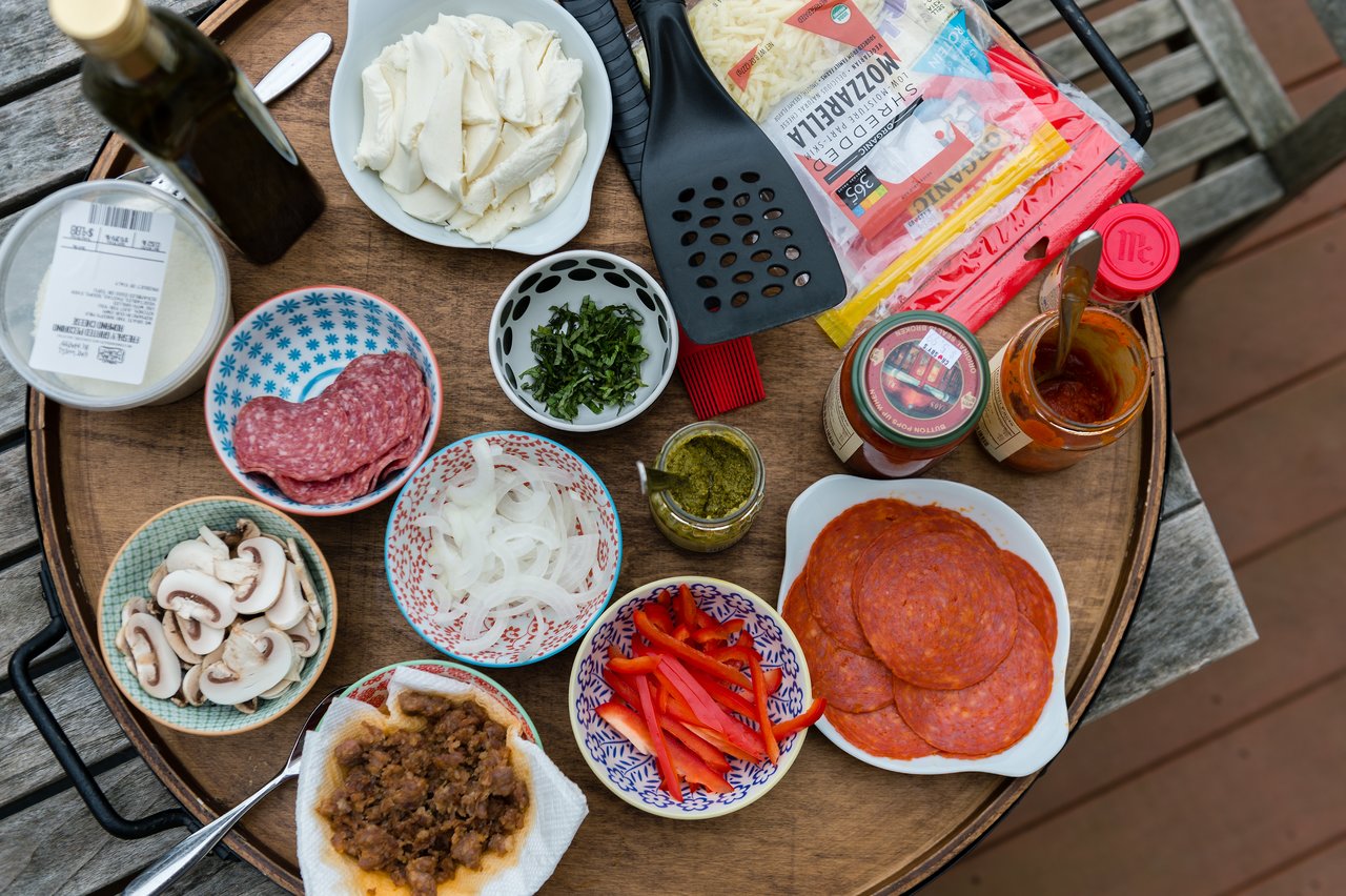 A round tray holds various pizza ingredients, including cheese, meats, vegetables, sauces, and utensils, ready for grilling.