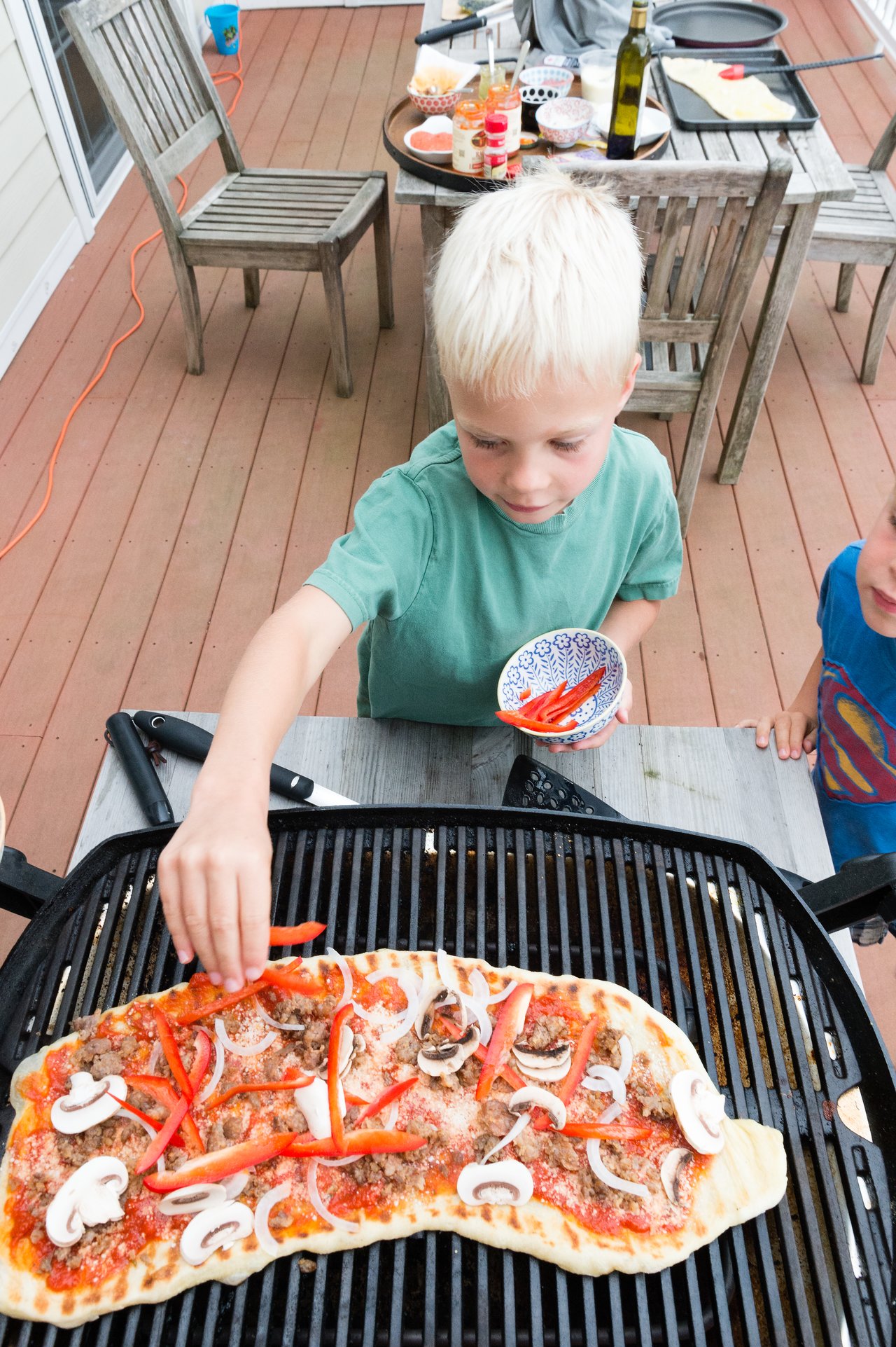 A child places red pepper slices on a homemade pizza cooking on a grill outdoors.