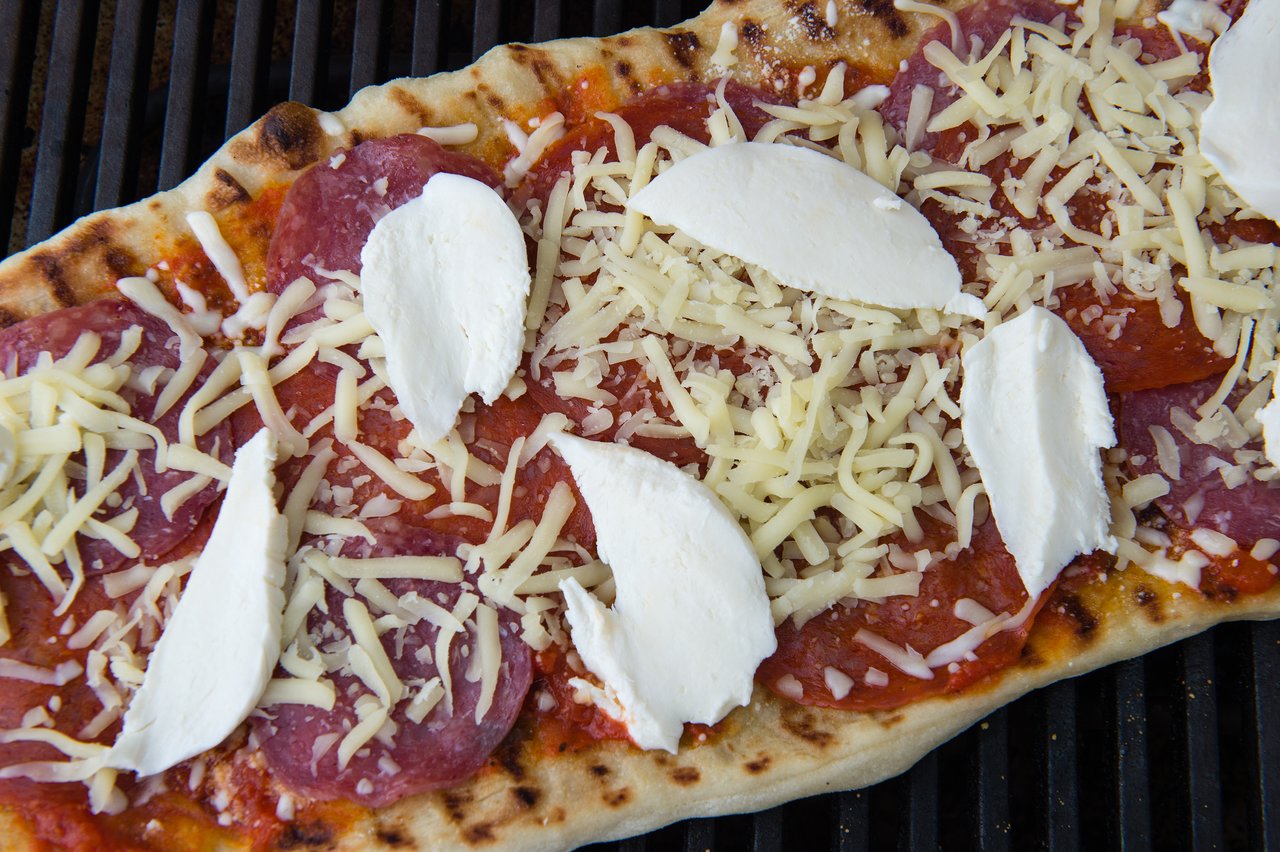Partially cooked pizza on a grill with tomato sauce, salami, shredded cheese, and fresh mozzarella slices.