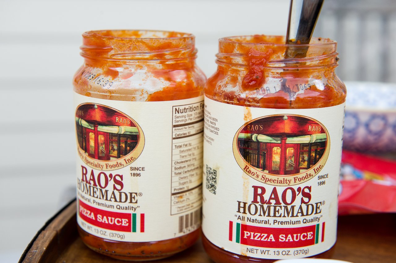 Two open jars of Rao's Homemade pizza sauce, one with a spoon inside, sitting on an outdoor surface.