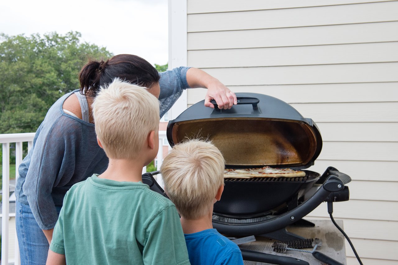 A woman lifts the lid of a grill while two children watch pizzas cooking on the grates.