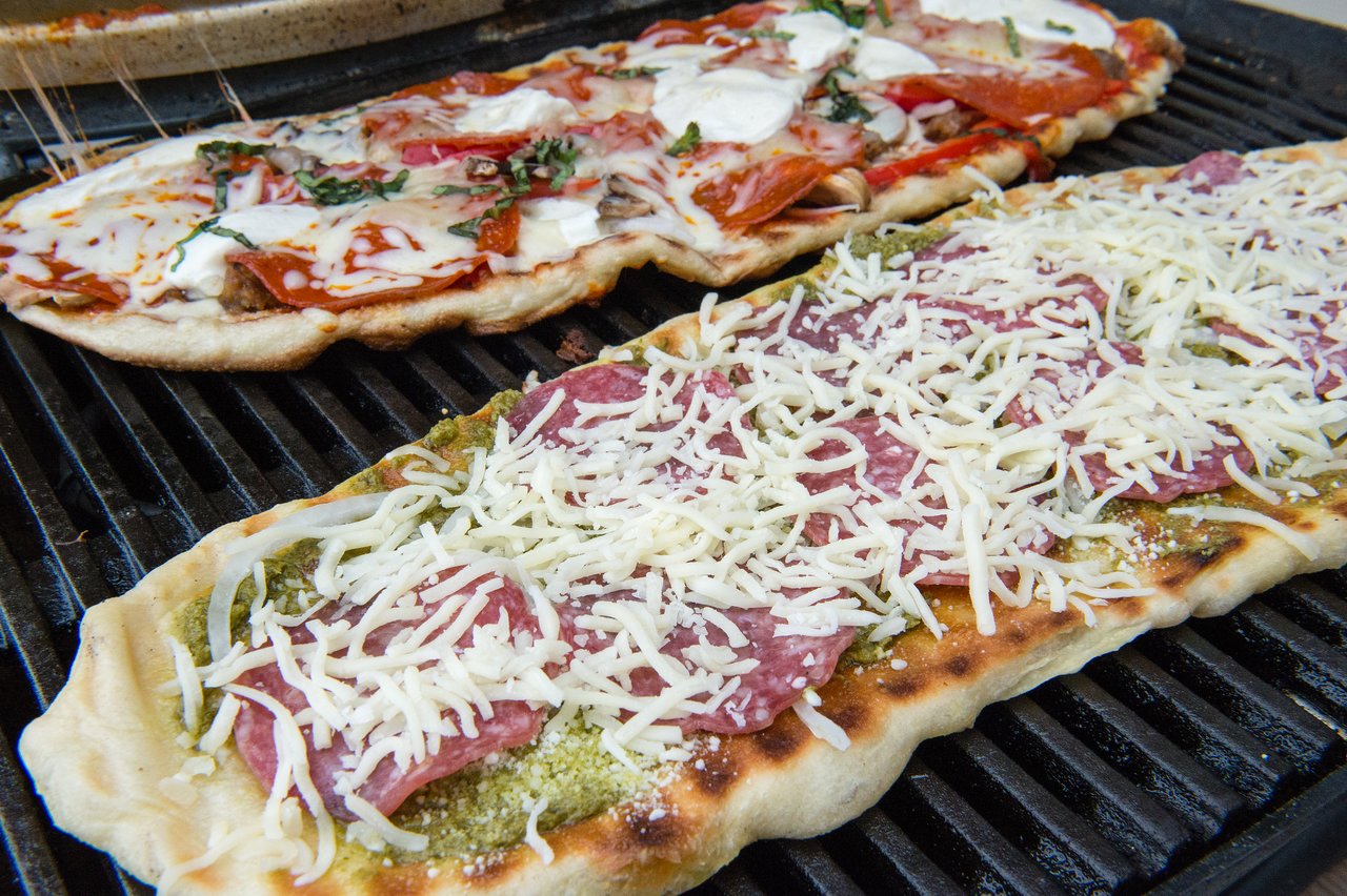 Two pizzas cooking on a grill, one with melted cheese and pepperoni, the other with pesto, salami, and shredded cheese.
