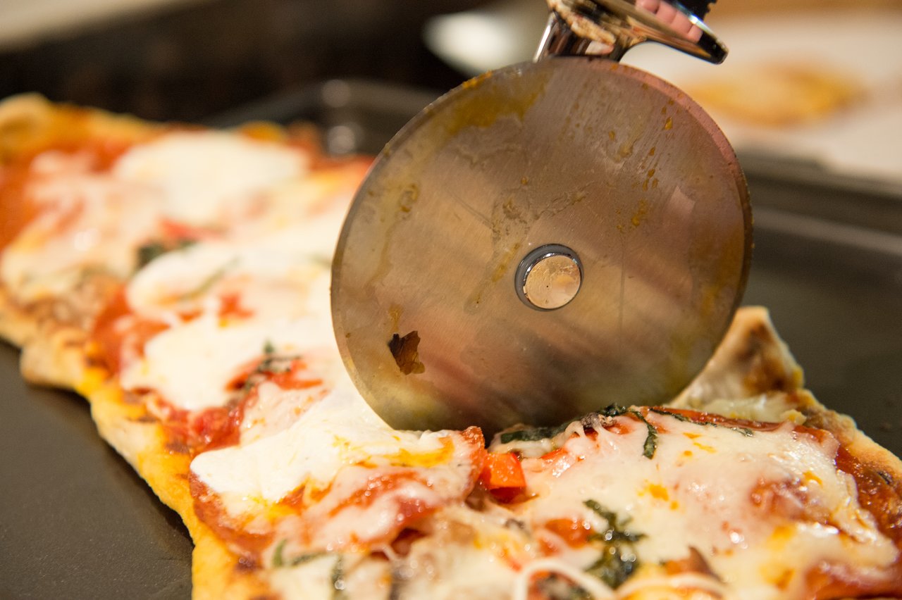 A pizza cutter slices a freshly grilled pizza with melted cheese and toppings on a tray.