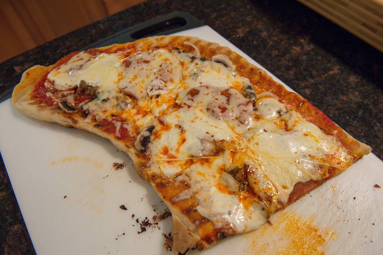 Grilled pizza with melted cheese, tomato sauce, and mushrooms on a cutting board, with a bite taken out.