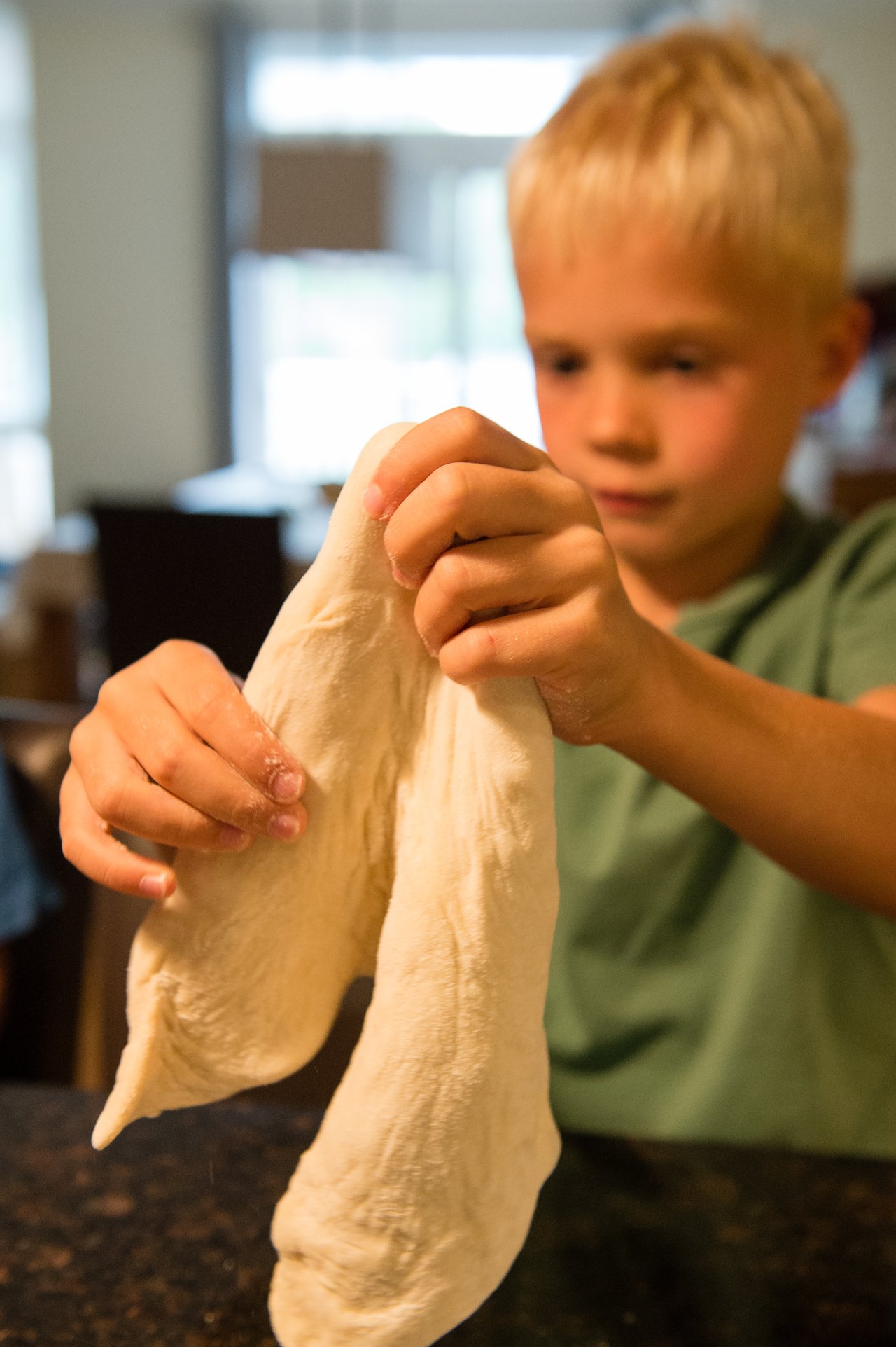 A child stretches pizza dough with both hands, preparing it for grilling.