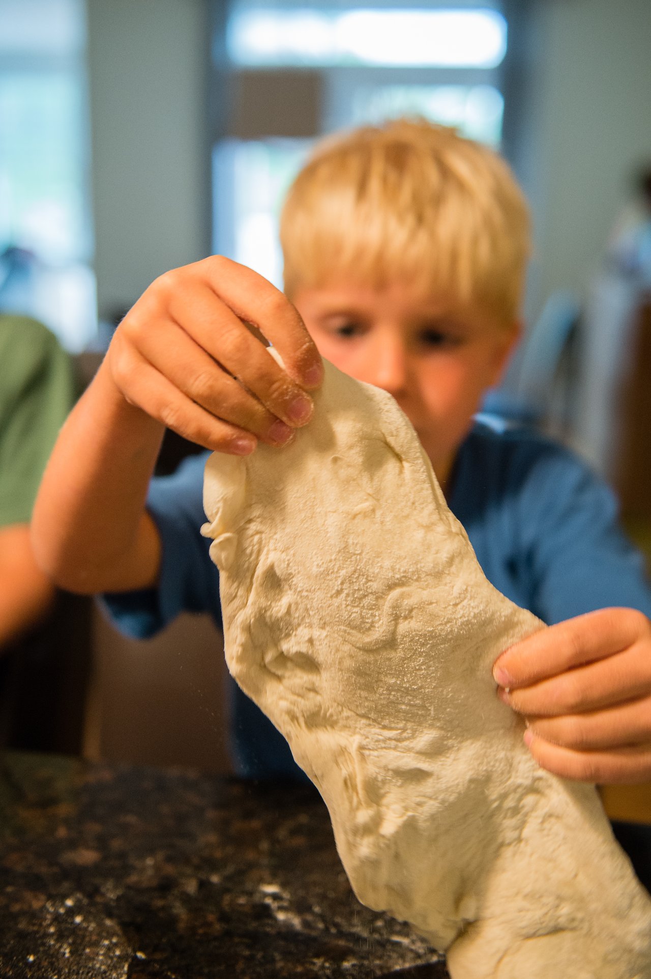 A child stretches pizza dough with both hands, preparing it for grilling.