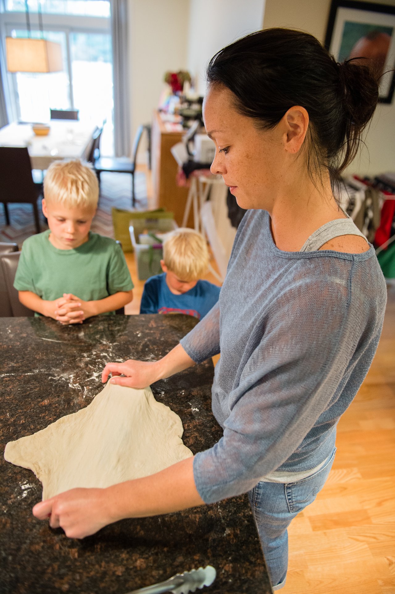 A woman stretches pizza dough on a kitchen counter while two children watch nearby.