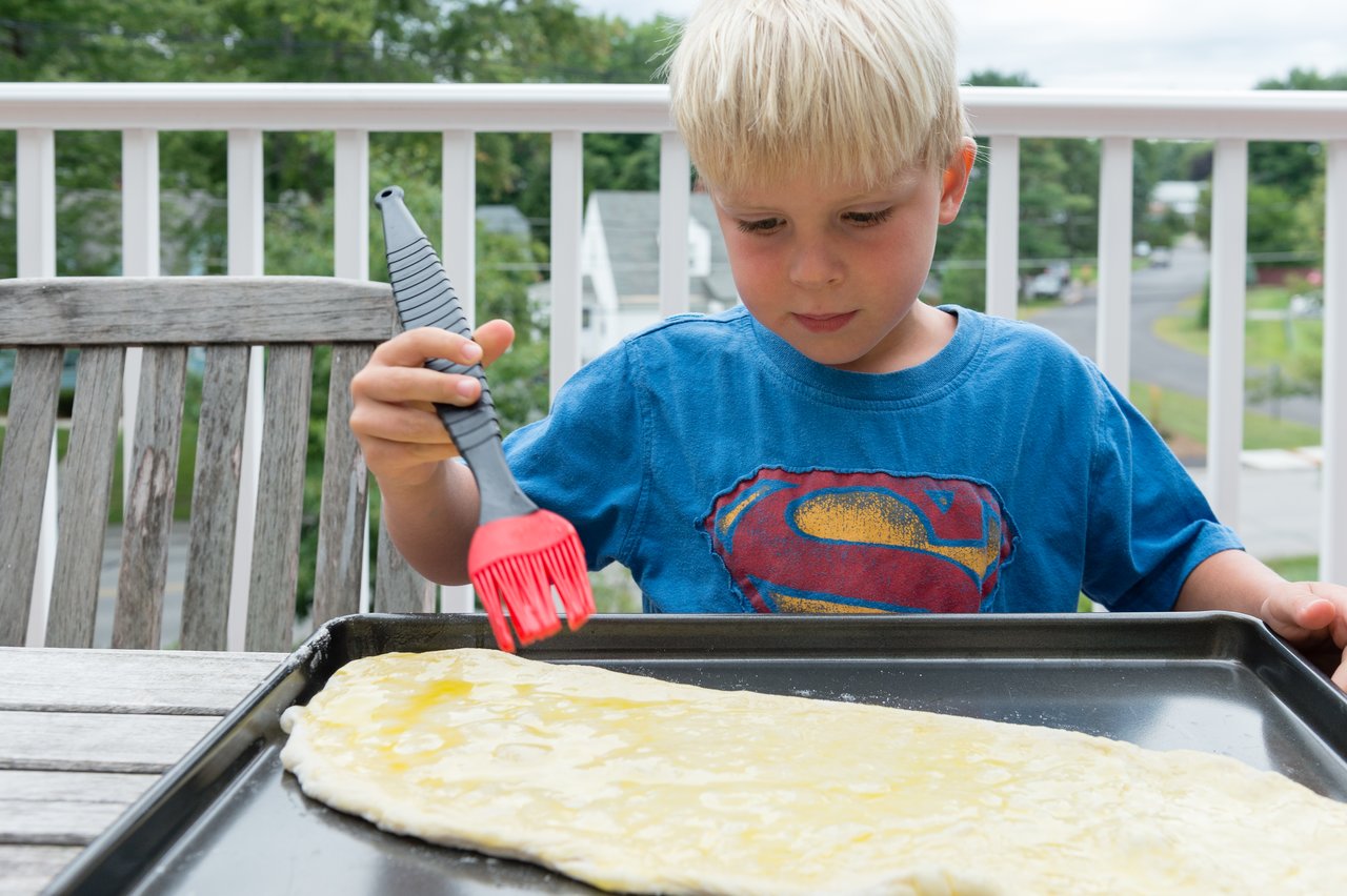 A child brushes oil onto pizza dough on a baking sheet, preparing it for grilling.