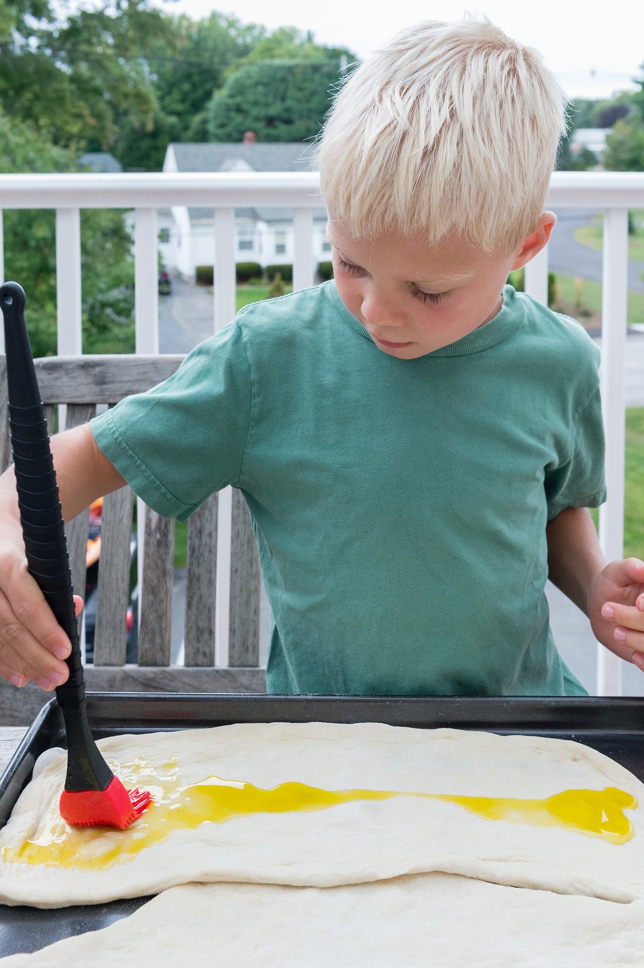 A child in a green shirt brushes oil onto pizza dough on a baking sheet outdoors.