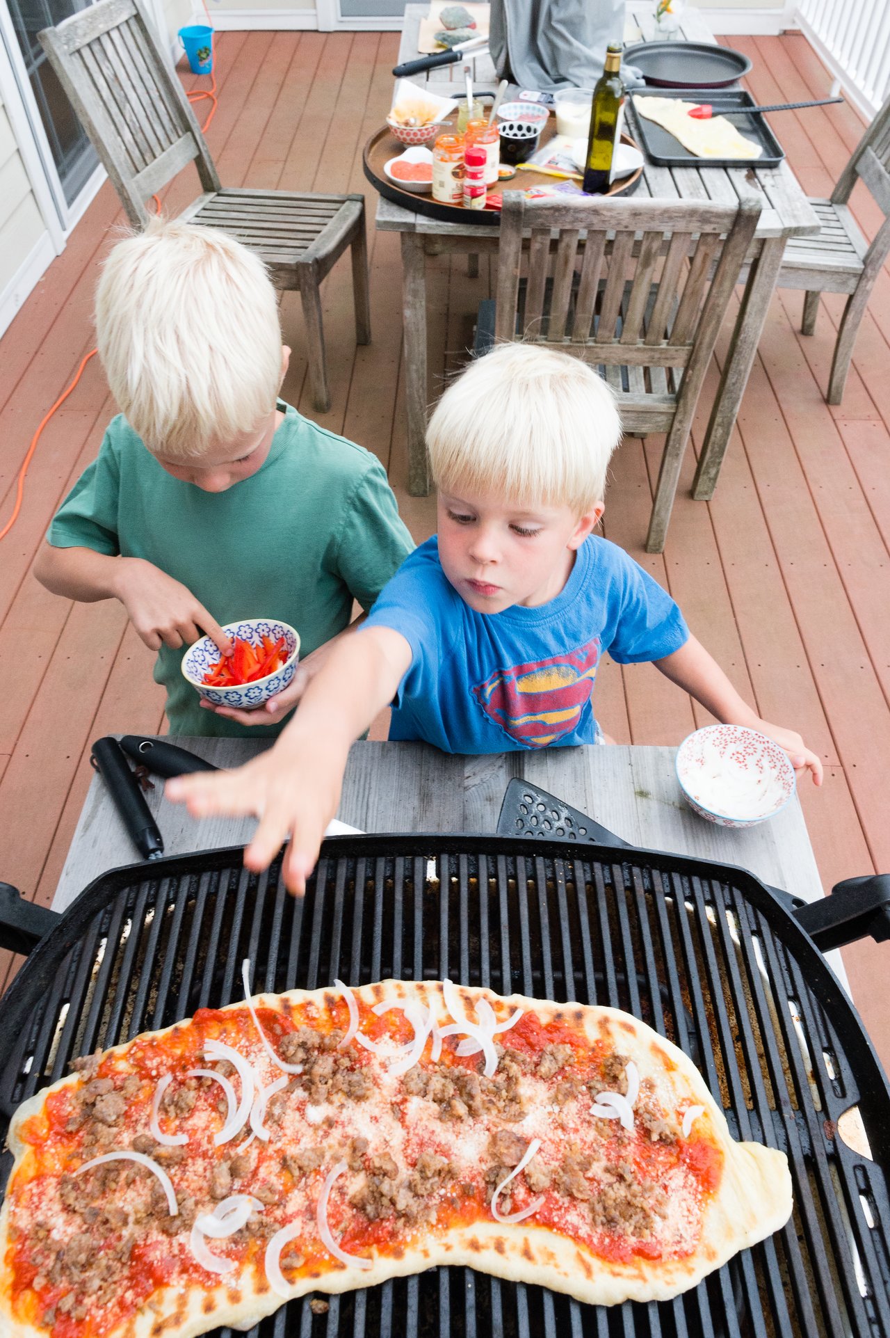 Two children prepare a homemade pizza on a grill, adding toppings like onions and sausage.