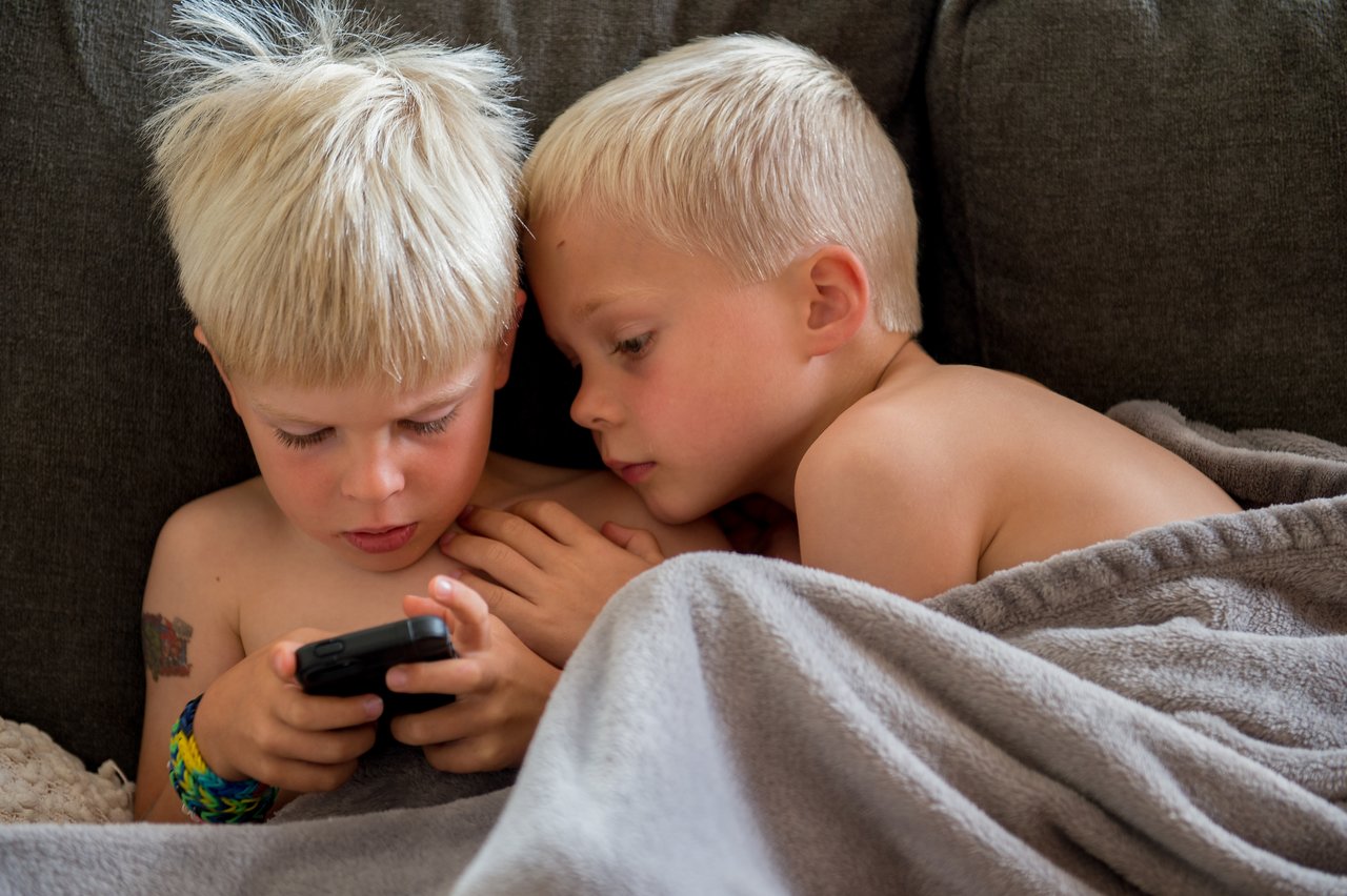 Two young boys sit closely on a couch, focused on a smartphone, with one wrapped in a blanket.