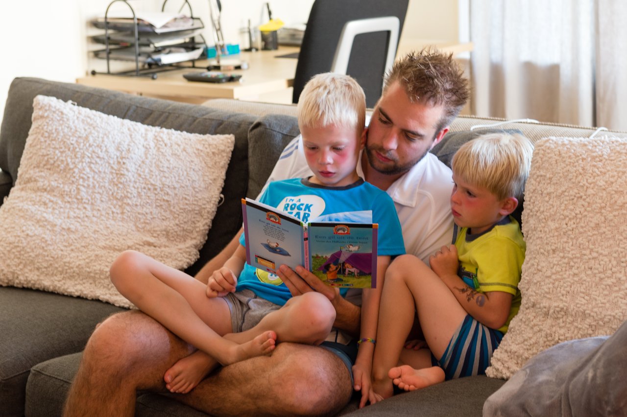 A man sits on a couch with two young boys, helping them read a children's book together.