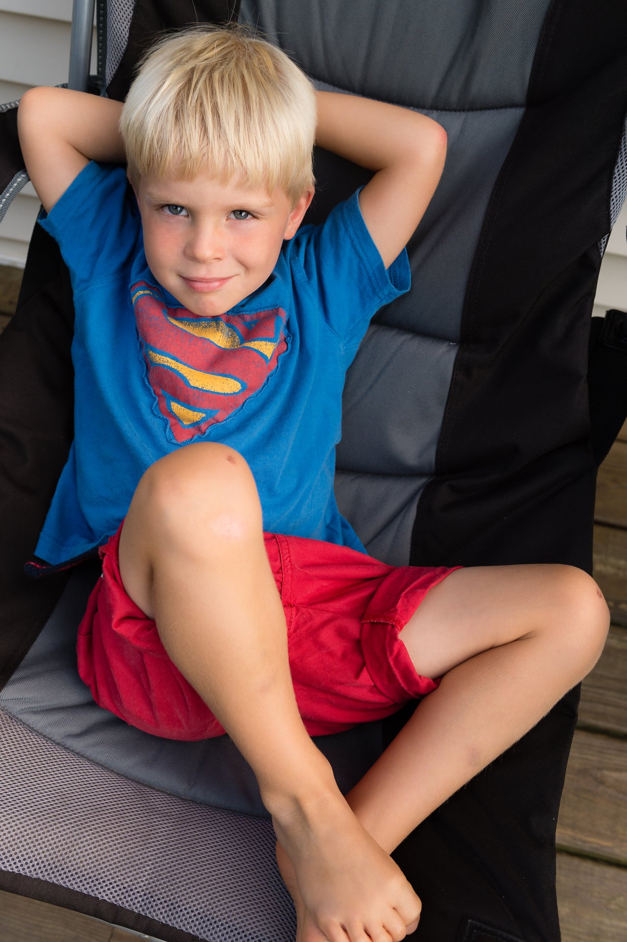 A young boy with blond hair sits in a chair, leaning back with his hands behind his head.