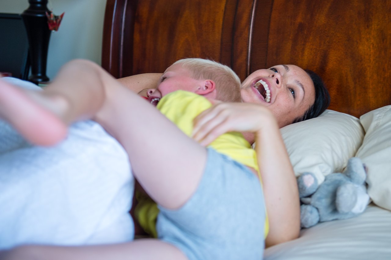 A woman and a young child laugh together on a bed while playfully tickling each other.