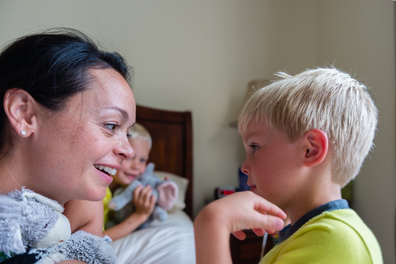 A woman smiles while playfully tickling a young boy, as another child watches from the bed holding a stuffed toy.