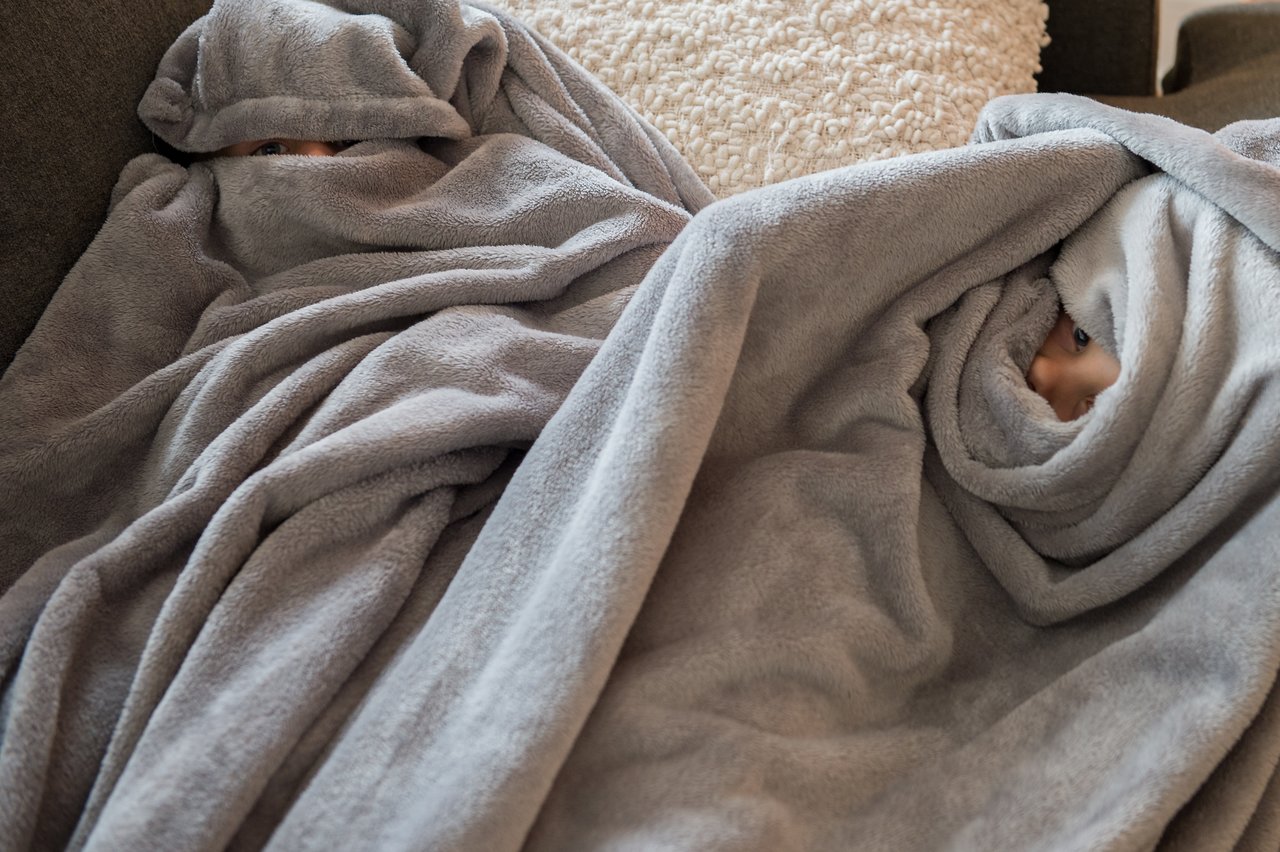 Two children are hiding under gray blankets on a couch, with only their eyes peeking out.