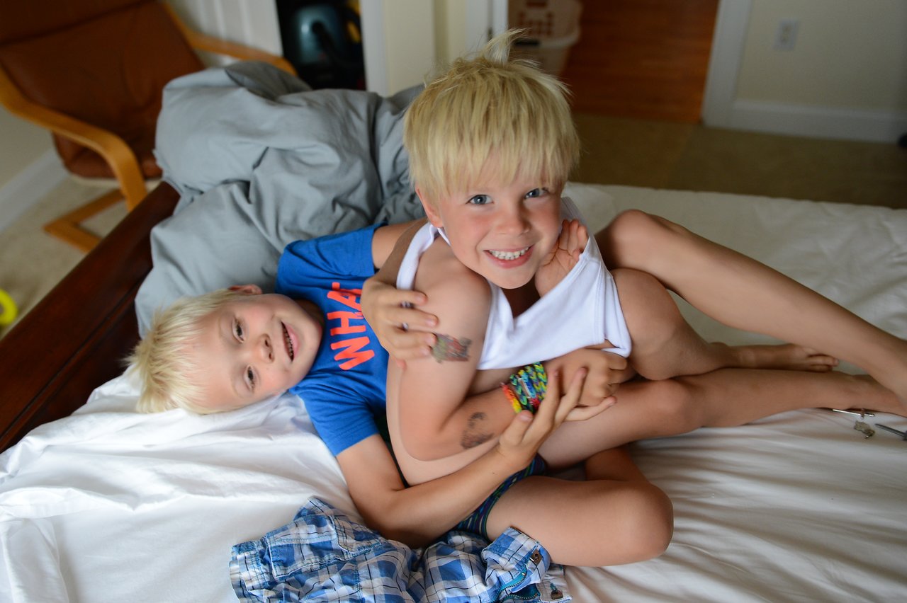 Two young boys with blonde hair playfully wrestle on a bed, smiling and laughing while holding onto each other.