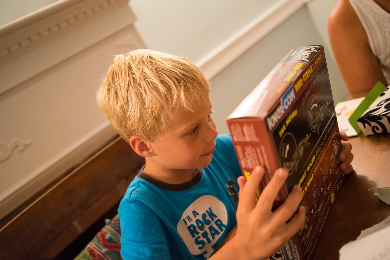 A young boy in a blue shirt excitedly examines a new toy box at his birthday party.