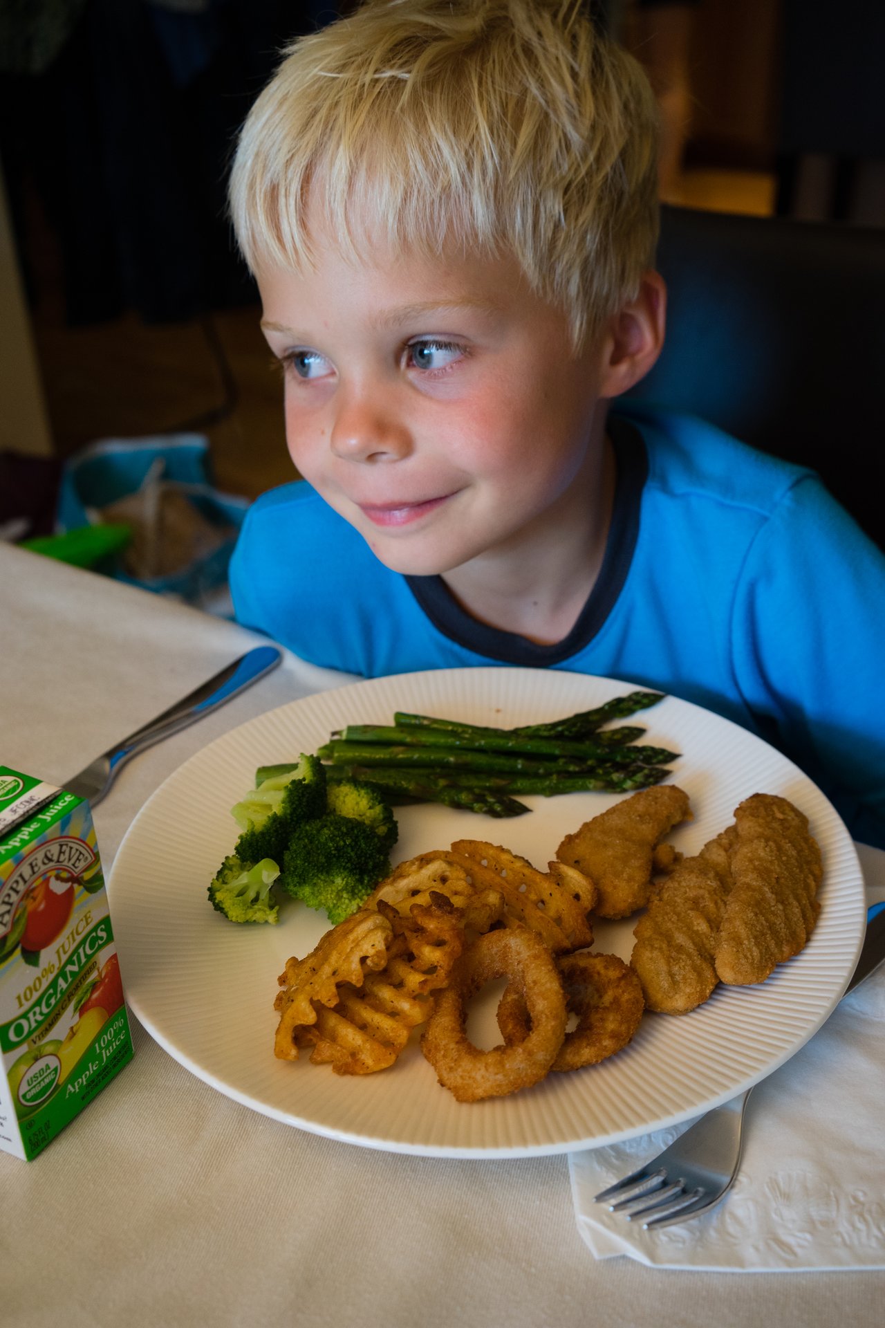 A young boy in a blue shirt sits at a table, smiling with a plate of food in front of him.