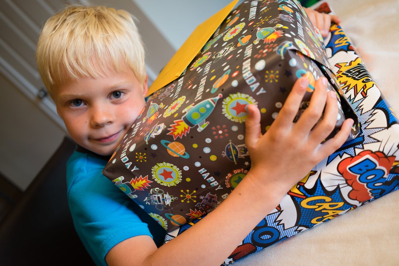 A young boy in a blue shirt holds a large, wrapped birthday present with colorful space and comic designs.