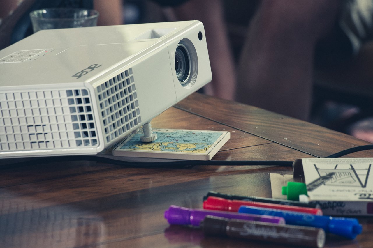 A projector rests on a wooden table with markers, a notebook, and a coaster underneath for support.