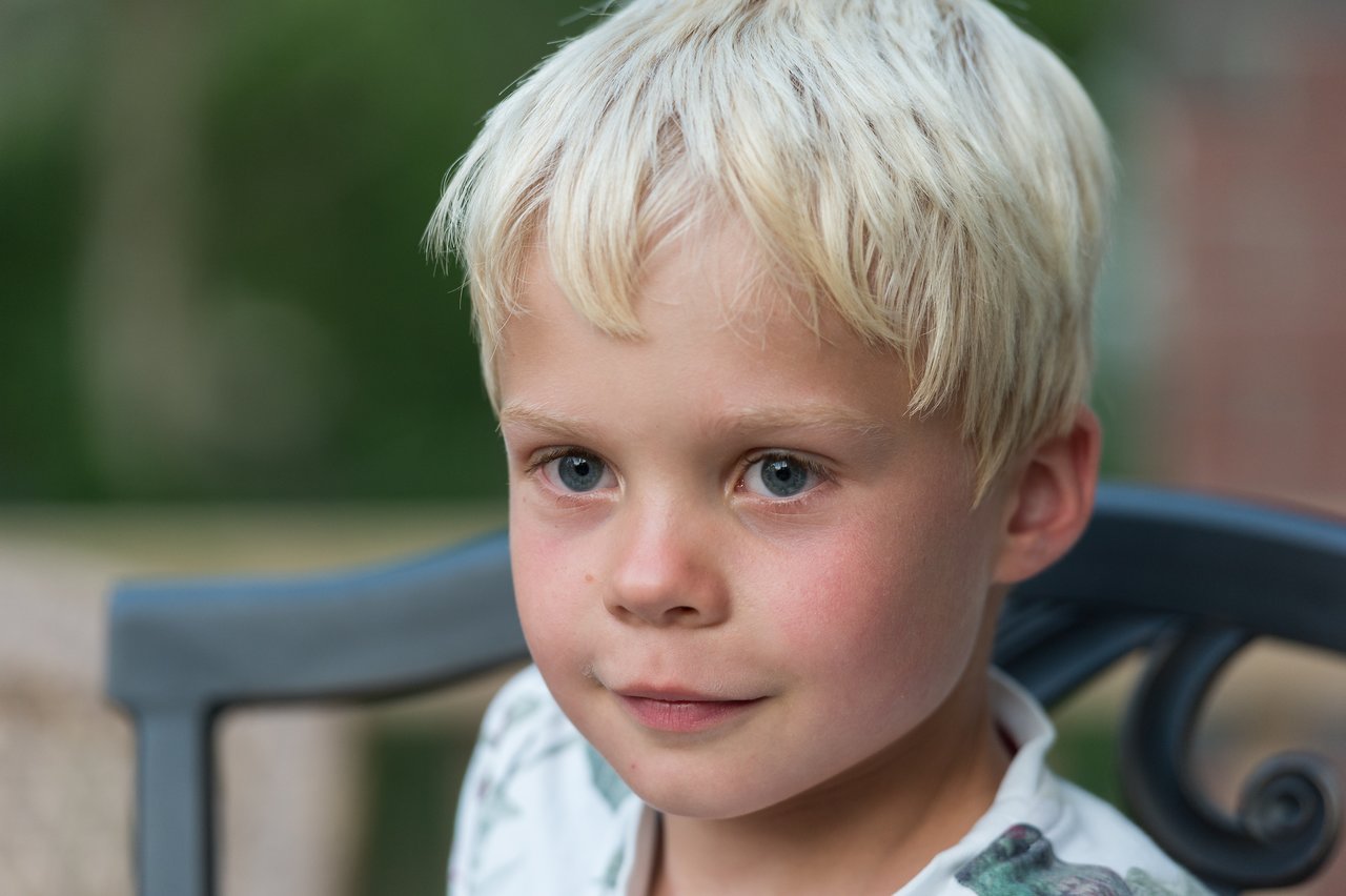 A young boy with blonde hair sits on a chair, looking directly at the camera with a calm expression.