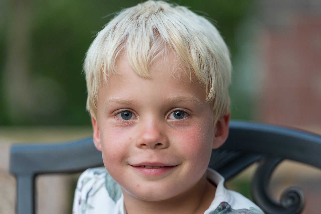 A young child with blonde hair sits on a metal chair, looking directly at the camera with a slight smile.