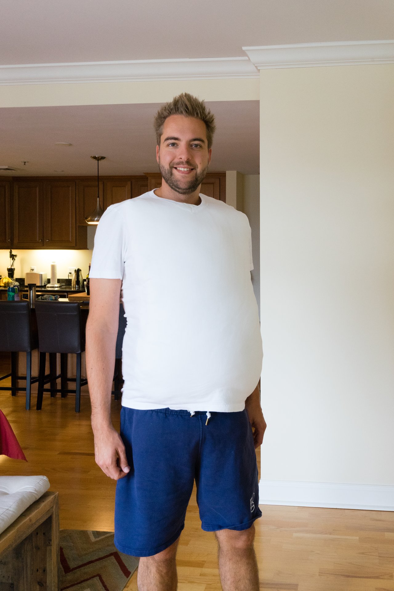 A man in a white shirt and blue shorts stands indoors, smiling with a noticeably rounded belly.