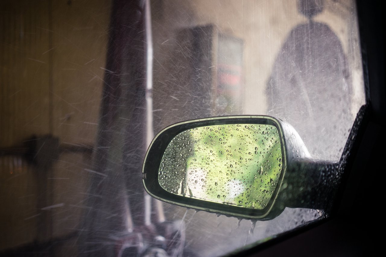Water sprays over a car's side mirror during a car wash, with droplets visible on the glass.