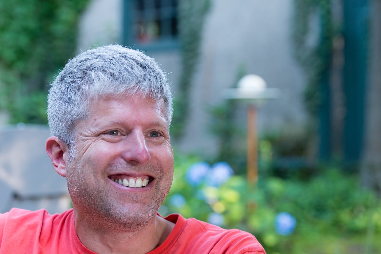 A man with short gray hair and a red shirt smiles while sitting outdoors in a garden setting.