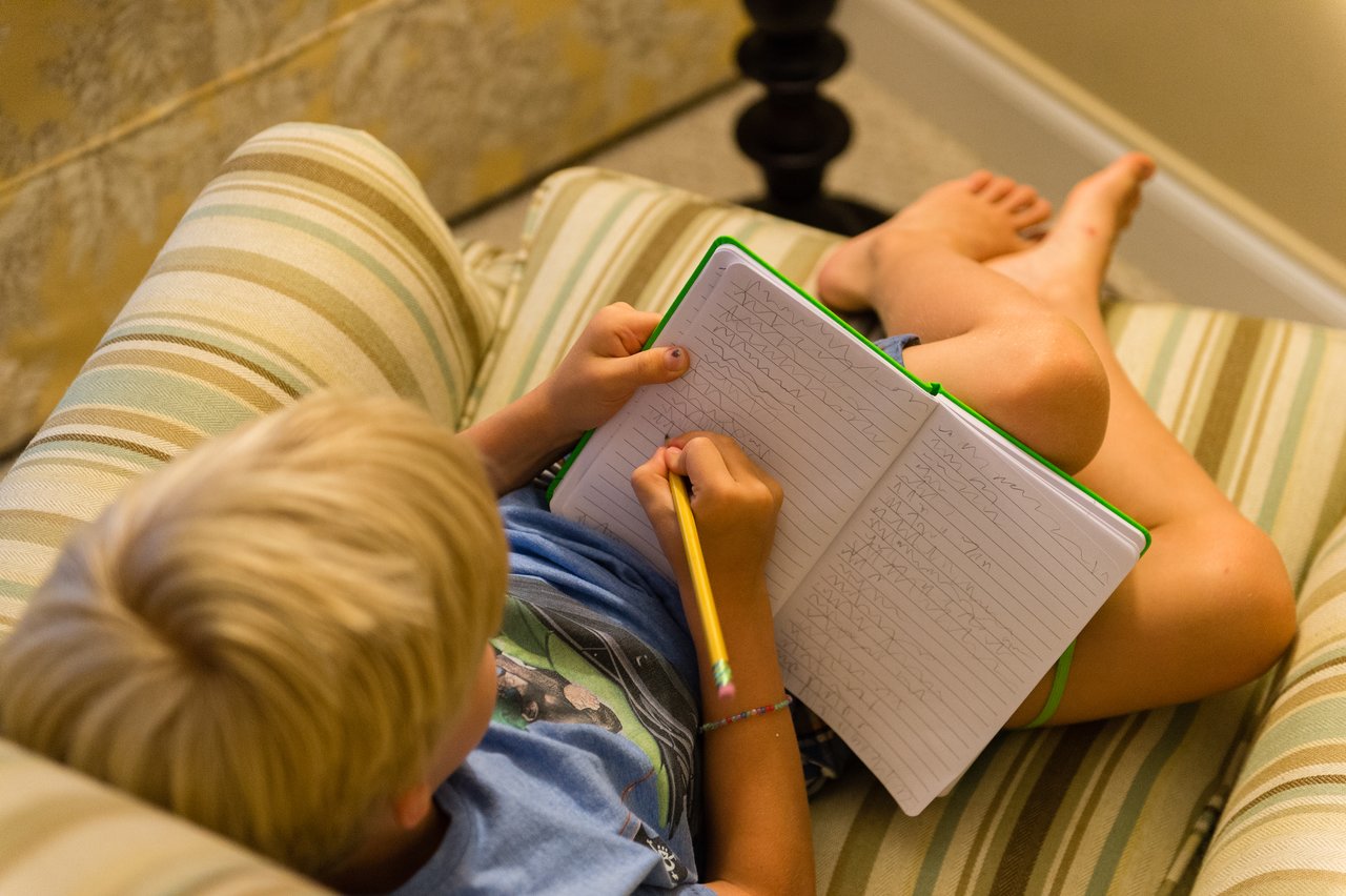 A child sits on a striped chair, writing in a notebook with a pencil.