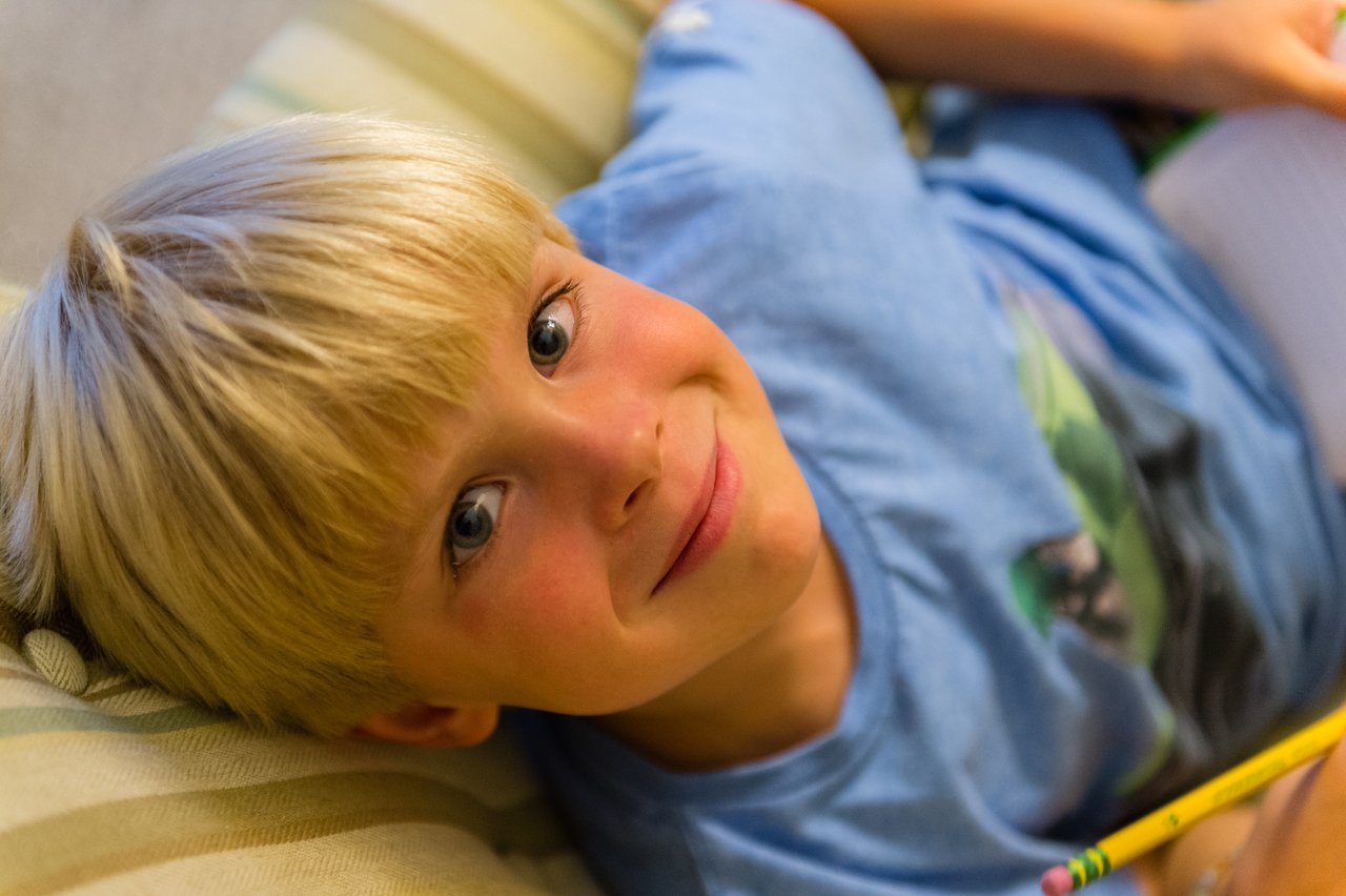 A child in a blue shirt smiles while holding a pencil and writing in a notebook.