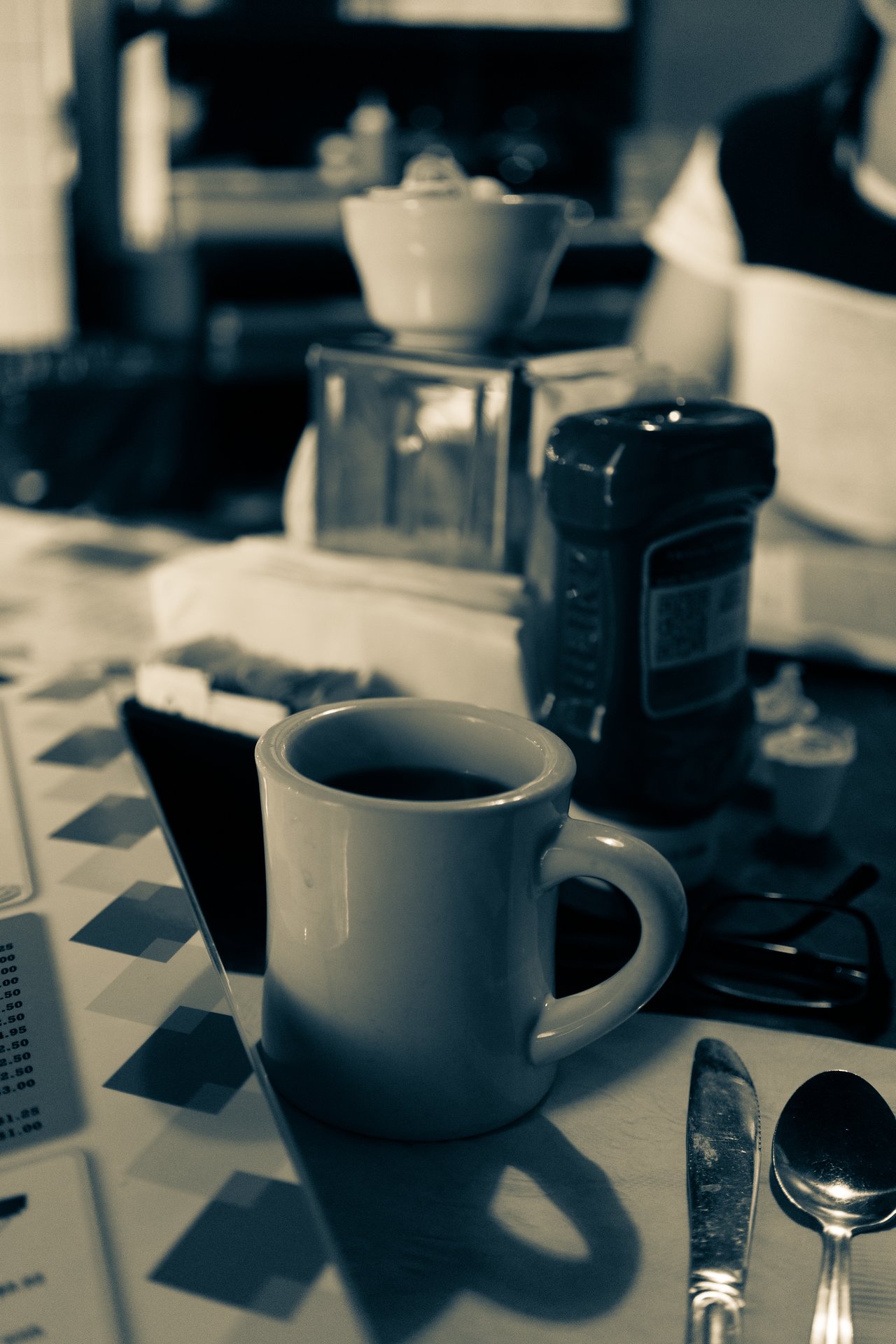 A cup of coffee sits on a diner table with a menu, utensils, and condiments nearby.