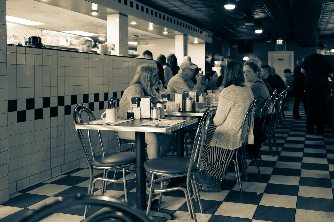 A group of people sits at a table in a busy diner, engaged in conversation and enjoying their meals.
