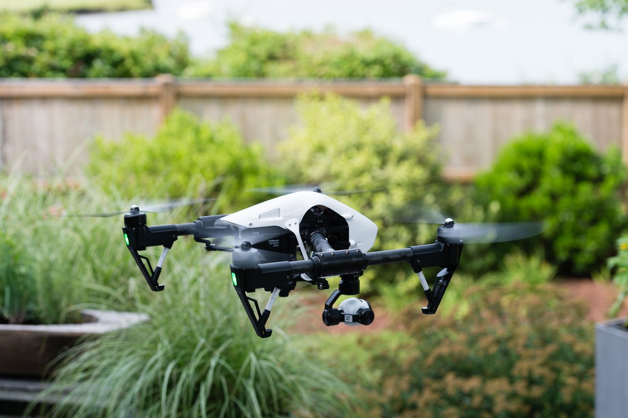 A white and black drone with a camera hovers in a backyard with greenery and a wooden fence.