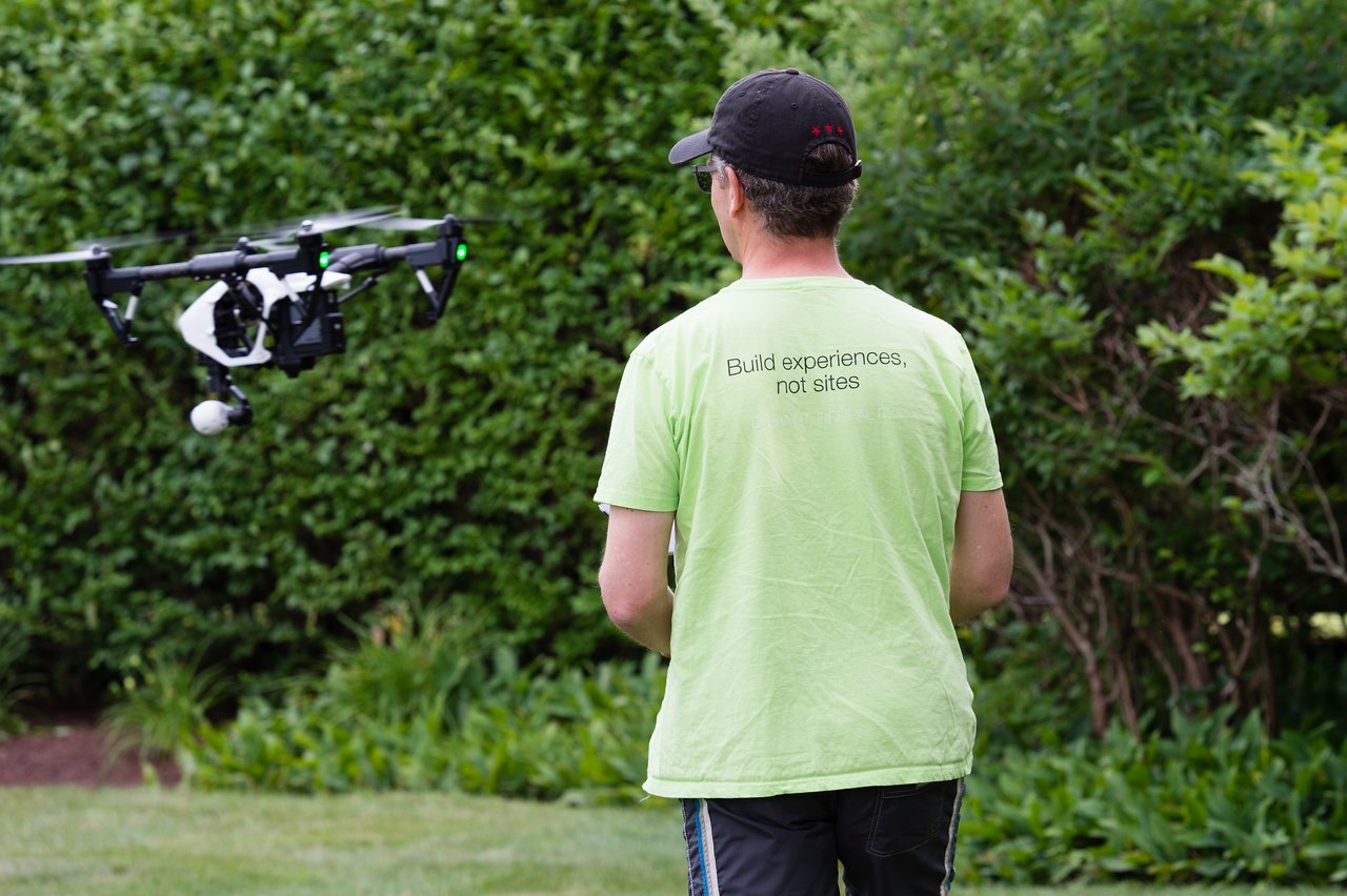 A person in a green shirt operates a drone hovering nearby in an outdoor setting with greenery.