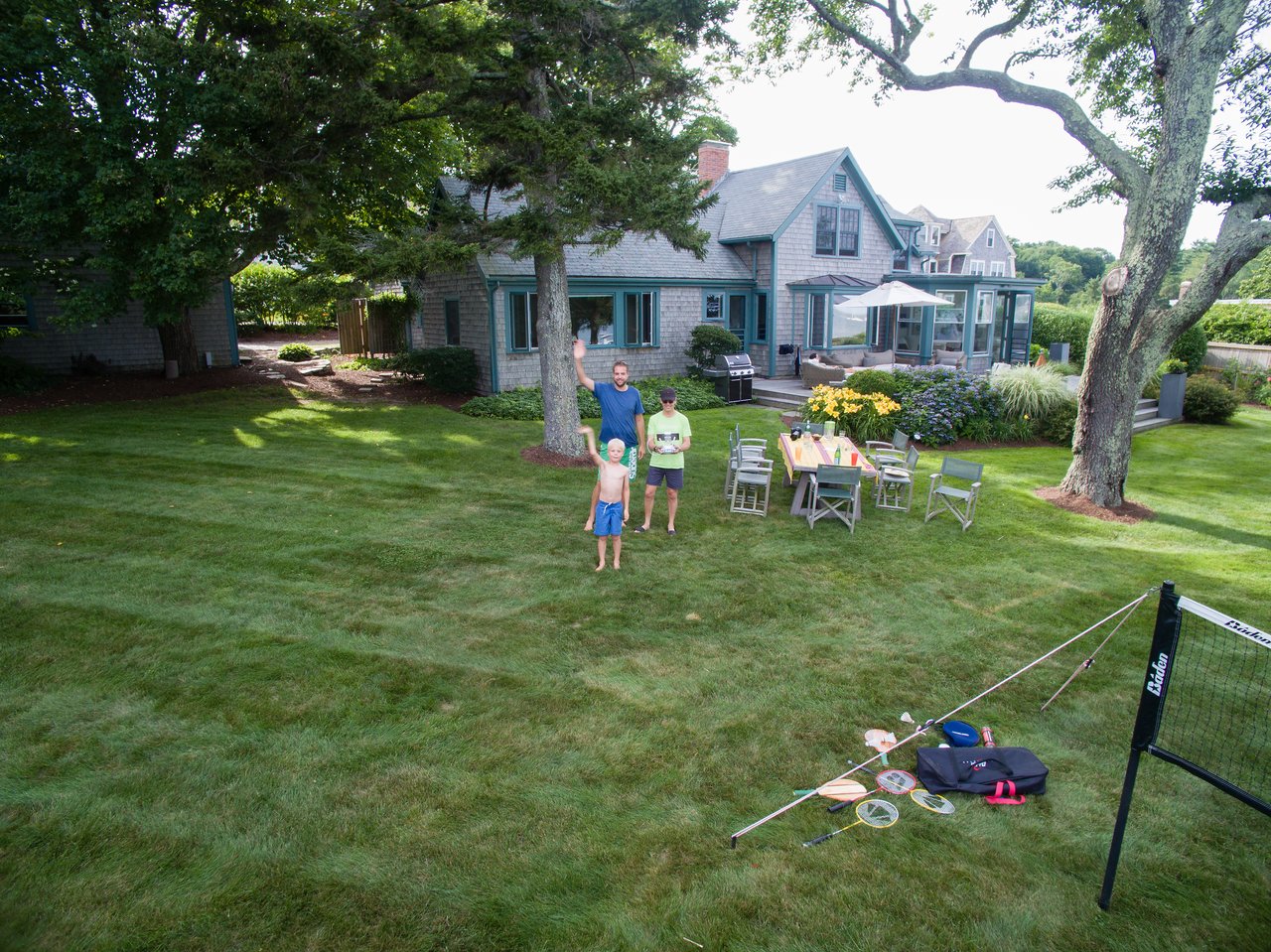Three people stand in a grassy backyard, one controlling a drone while two wave at the camera.