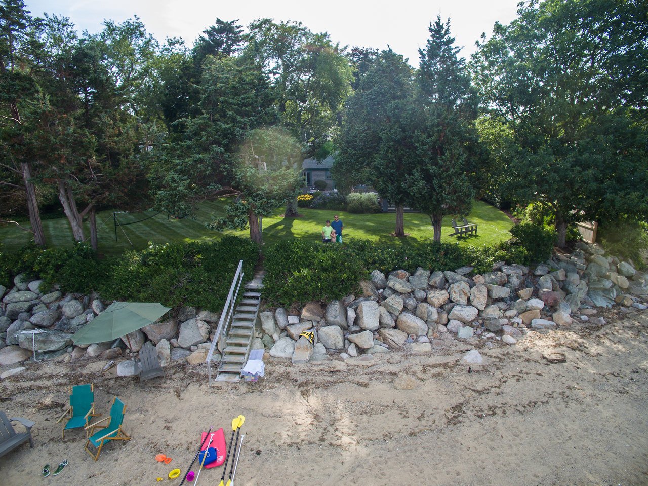 Two people stand on a grassy hill, looking up at a drone capturing the photo near a beach.