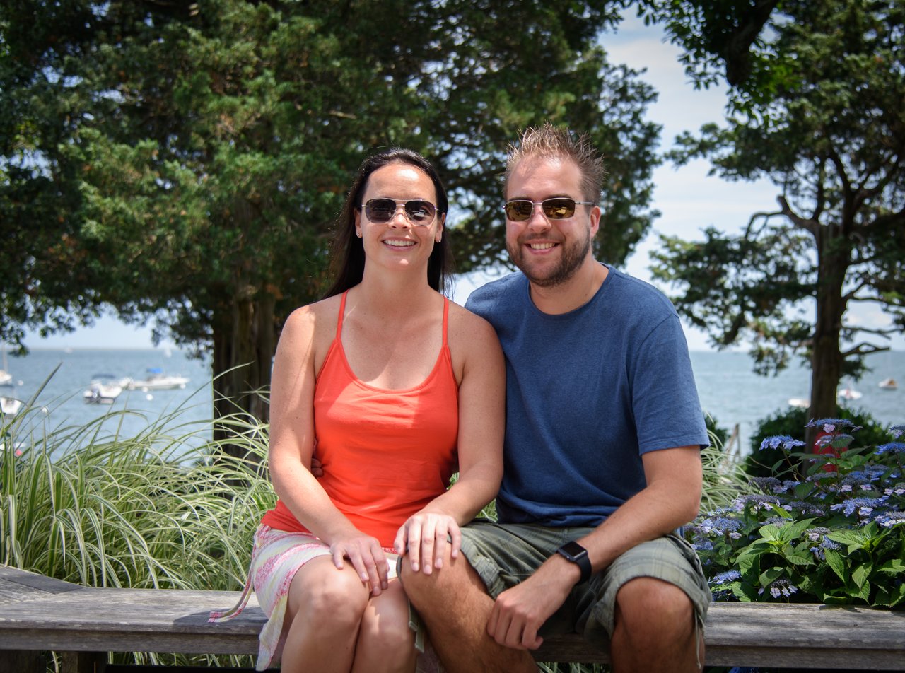 A smiling couple wearing sunglasses sits on a bench with a scenic waterfront and boats in the background.