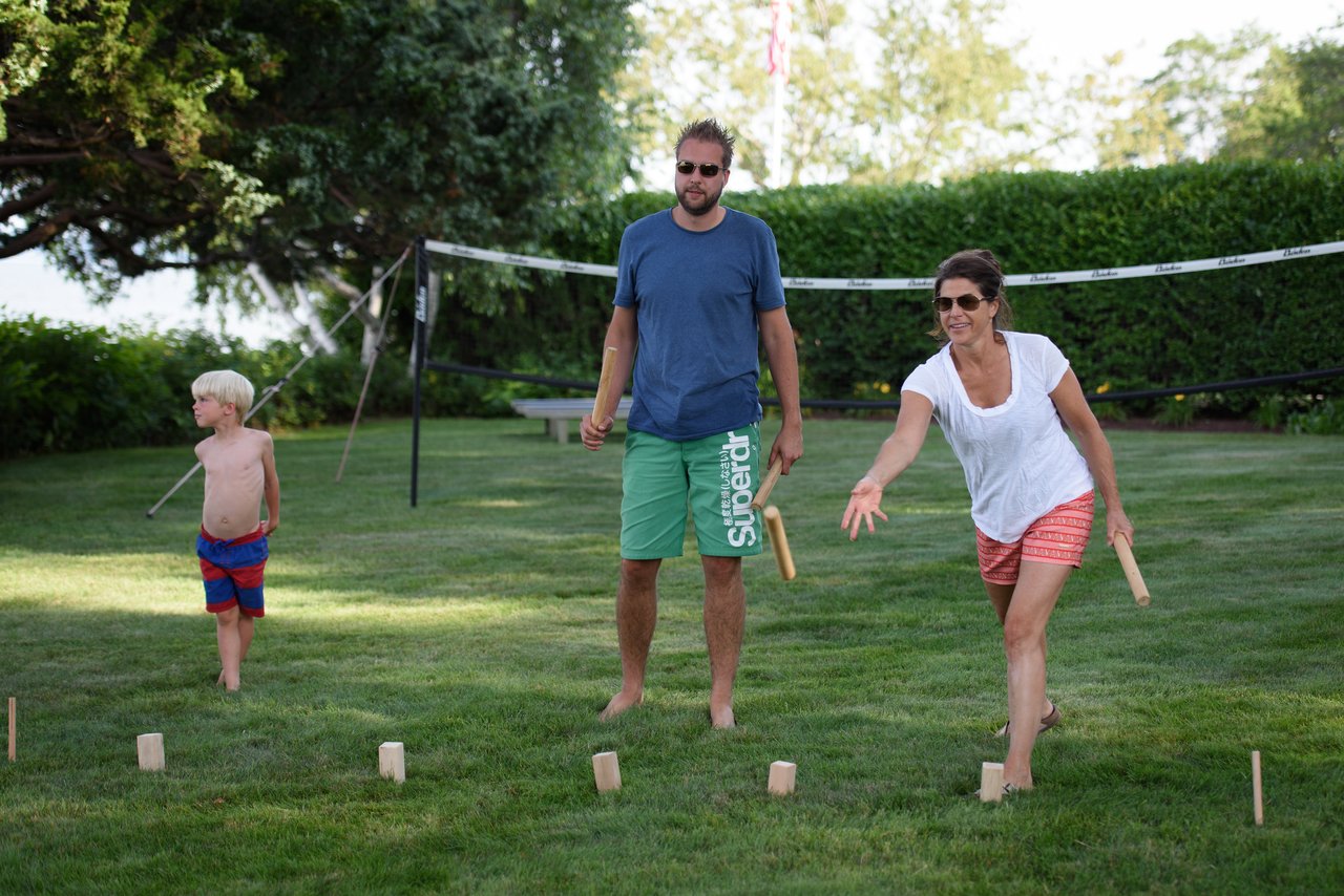 A woman throws a wooden stick while playing a lawn game, with a man and child watching nearby.