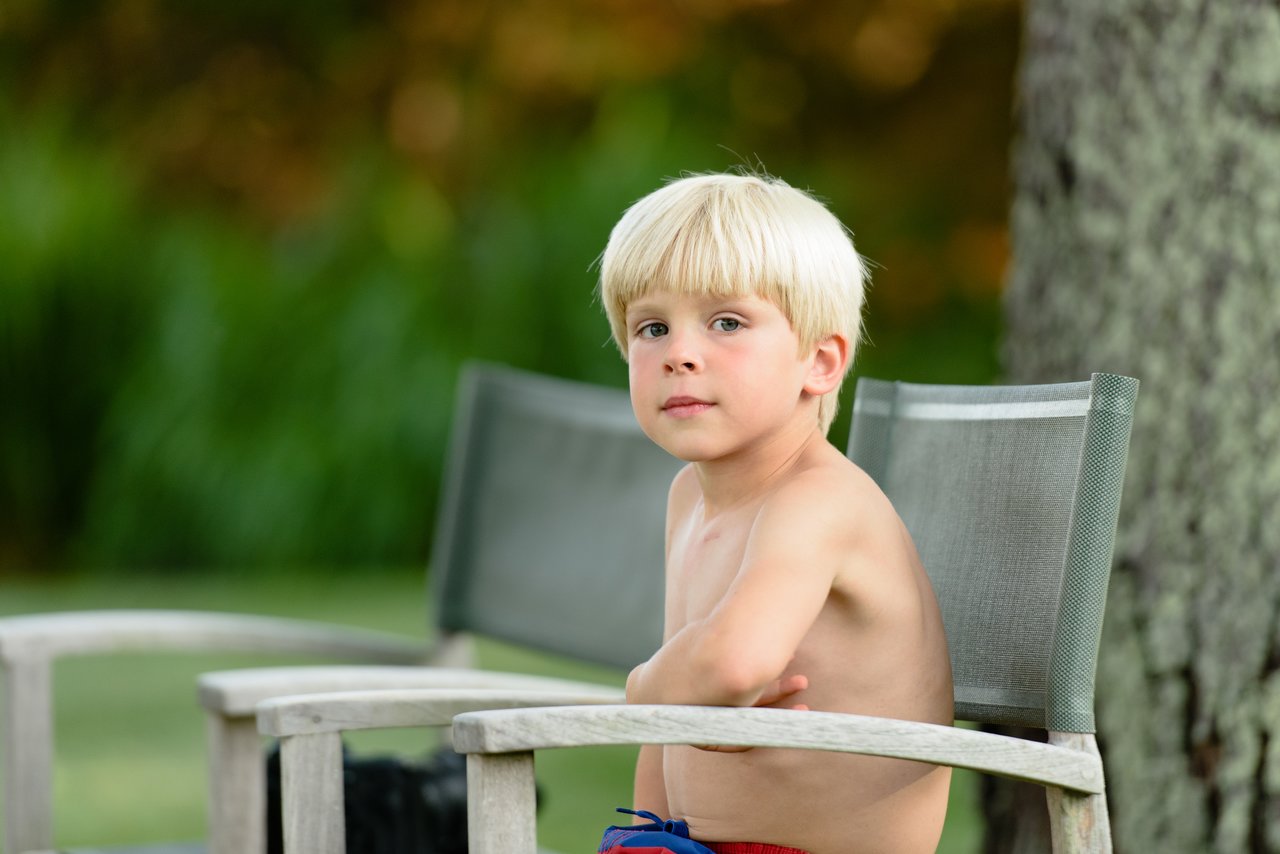A young boy with blond hair sits shirtless on a patio chair, looking toward the camera.