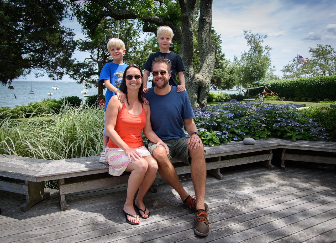 A family of four poses on a wooden bench in a garden with a view of the water.