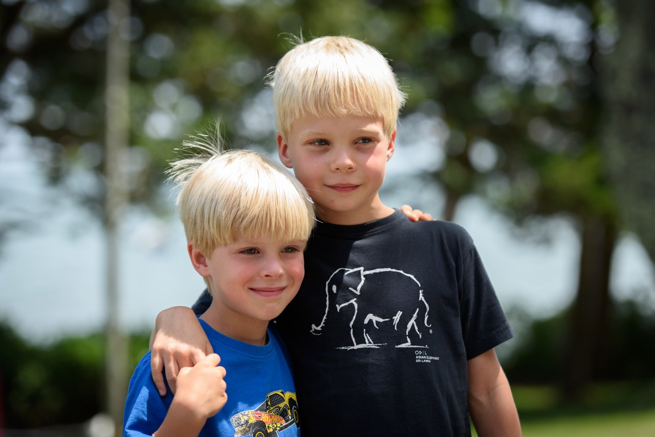 Two young boys stand close together, smiling, with one wrapping his arm around the other in an outdoor setting.
